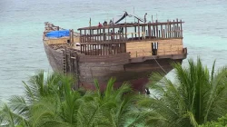Lambung kapal kayu besar yang sedang dibangun di tepi pantai berair biru kehijauan, beberapa pekerja berdiri di geladak, dengan daun kelapa di latar depan