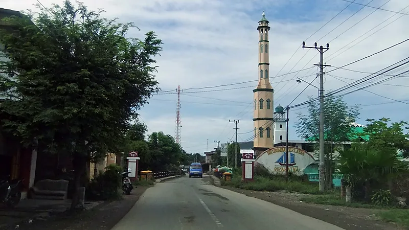 3.054 PPPK Paruh Waktu Bulukumba Dapat Penghasilan Minimal Rp750 Ribu Jalan beraspal di Bulukumba dengan menara masjid tinggi dan papan penanda bergambar perahu pinisi
