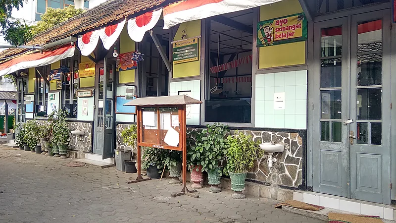 Teras depan gedung sekolah dasar berdinding kuning dengan pot tanaman, papan pengumuman kayu, dan hiasan merah putih
