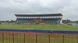 Tribun utama Stadion Sultan Agung beratap baja dengan kursi biru dan merah, sementara dua tim bertanding di lapangan hijau di balik pagar besi