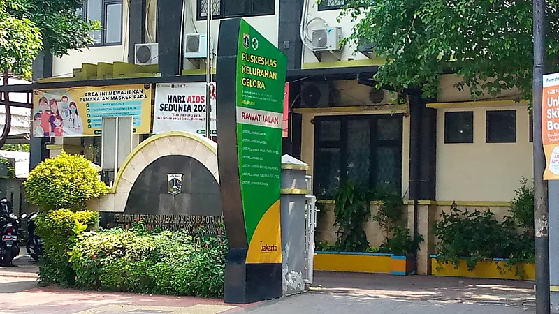 21 Puskesmas di Bulukumba dan Sebarannya per Kecamatan Papan penanda tegak berwarna hijau bertuliskan Puskesmas Kelurahan Gelora dan Rawat Jalan berdiri di trotoar di depan gedung berpagar, dengan spanduk layanan kesehatan terpasang di dinding