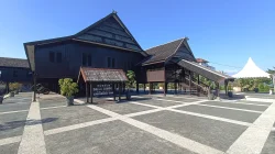 Bangunan kayu panggung bergaya rumah adat Bugis-Makassar bercat gelap dengan papan bertuliskan Museum Balla Lompo Sungguminasa - Gowa, berdiri di atas pelataran berpetak di bawah langit biru