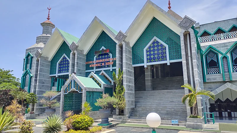 Fasad samping Masjid Al-Markaz Al-Islami dengan tiga gevel runcing berlapis ornamen kisi hijau toska, papan nama berhuruf merah di dinding, tangga lebar menuju pintu masuk, dan taman kecil berpot di depannya