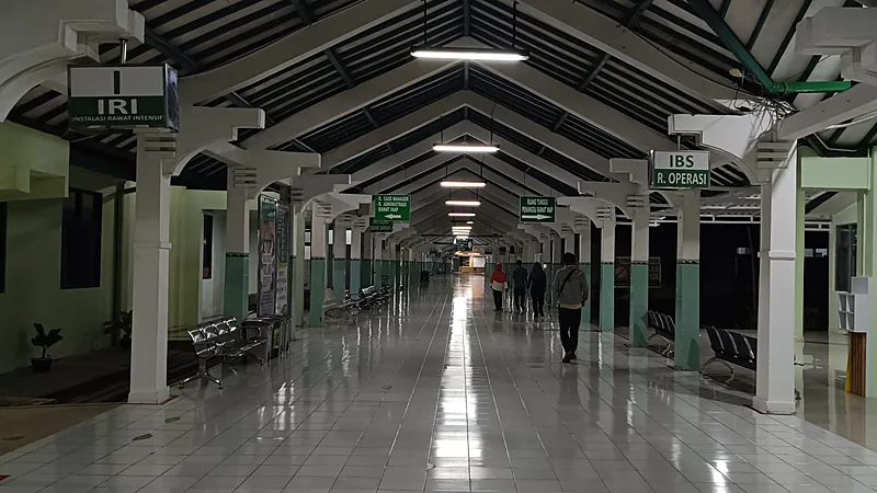 RSUD Andi Sulthan Daeng Radja Bulukumba: Kelas B, Paripurna Lorong panjang rumah sakit beratap rangka kayu dengan lantai keramik mengilap, papan penunjuk instalasi rawat intensif dan ruang operasi, serta beberapa orang berjalan