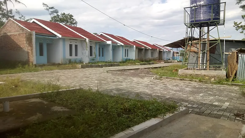 Jalan paving di antara deretan rumah subsidi berdinding biru putih, dengan menara tandon air biru dan bangunan berdinding seng di sisi kanan
