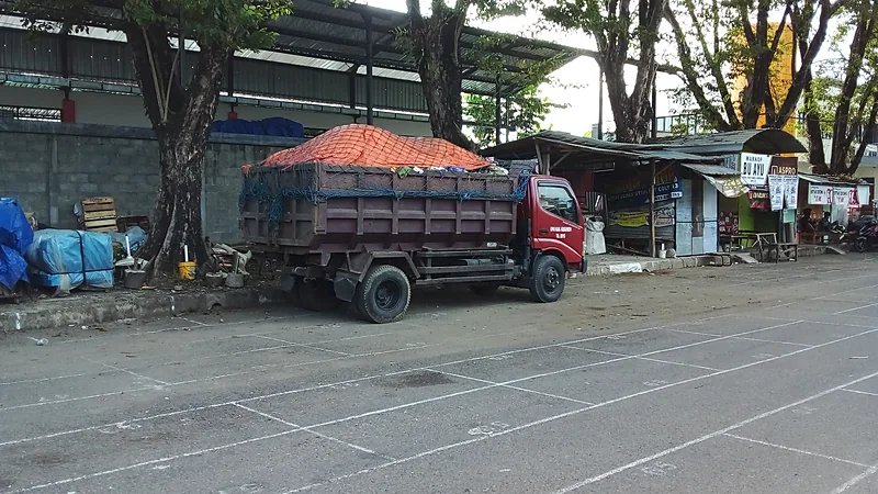 Sampah Menumpuk di Gowa, DPRD Panggil DLH dan Pemilik TPA Truk sampah bak terbuka berwarna merah dan cokelat dengan muatan tertutup terpal jaring oranye terparkir di tepi lapangan beraspal bergaris putih, dengan pepohonan dan deretan kios di belakangnya