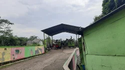 Jalan berpaving menuju tempat pengolahan sampah beratap seng dengan beberapa kontainer sampah hijau dan kendaraan pengangkut di bawahnya, diapit tembok bermural warna-warni di kiri dan dinding hijau di kanan, di bawah langit mendung