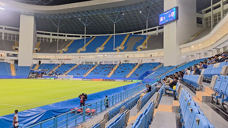 Interior Stadion Batakan dengan tribun berkursi biru dan penonton di sisi lapangan saat laga malam