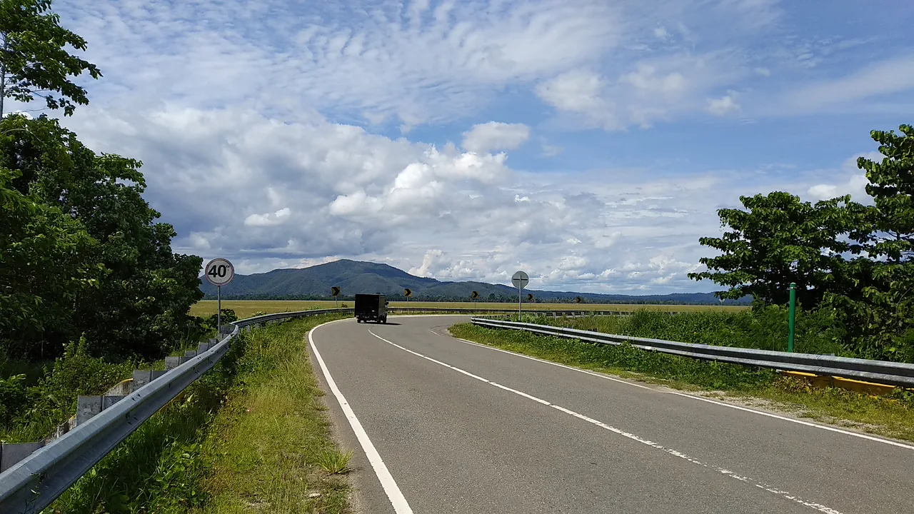 Jalan aspal dua lajur bermarka putih menikung di antara pagar pengaman dan rerumputan