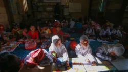 Anak-anak berseragam sekolah dasar duduk dan menulis di atas tikar di ruang berdinding papan kayu, dengan rak buku dan poster di dinding