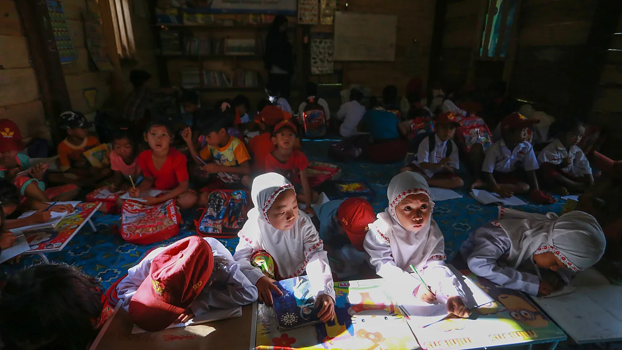 Anak-anak berseragam sekolah dasar duduk dan menulis di atas tikar di ruang berdinding papan kayu, dengan rak buku dan poster di dinding