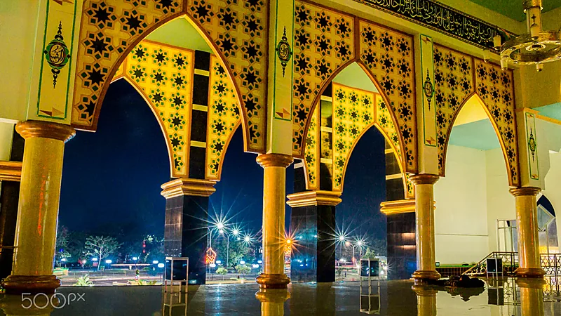 Sejarah Islam di Bulukumba dan Makam Dato Tiro Serambi masjid bernaung deretan lengkung runcing berhias mozaik bintang kuning keemasan dan kaligrafi, tiang-tiang bulat memantul di lantai marmer yang basah, dengan lampu jalan menyala di kegelapan malam di luar