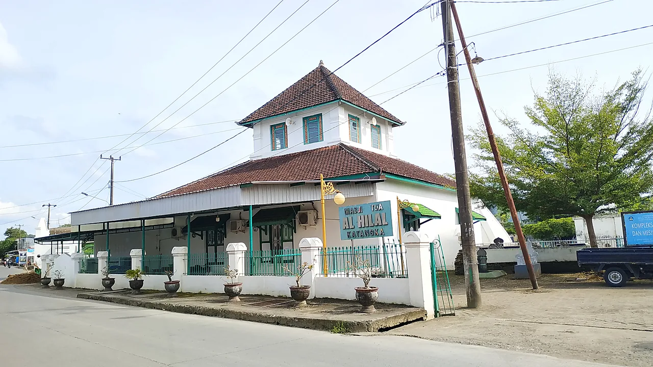Masjid tua beratap tumpang dua tingkat berdinding putih dengan pagar besi hijau di tepi jalan, papan bertuliskan Masjid Tua Al-Hilal Katangka terpasang di sisi depan