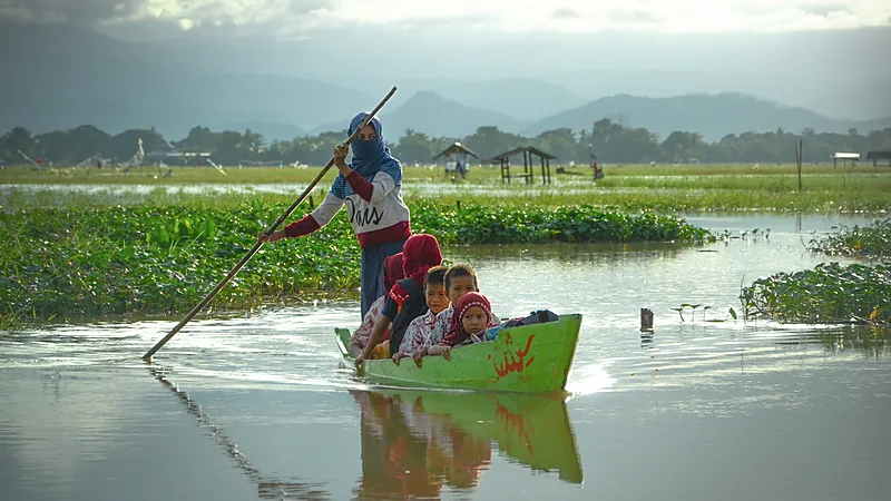 Sekolah Rakyat Sinjai Tuntas, Bulukumba Belum Masuk Daftar Seorang perempuan berjilbab mengayuh perahu kayu kecil berwarna hijau yang mengangkut beberapa anak berseragam merah putih melintasi perairan dangkal berumpun eceng gondok, dengan barisan bukit di kejauhan