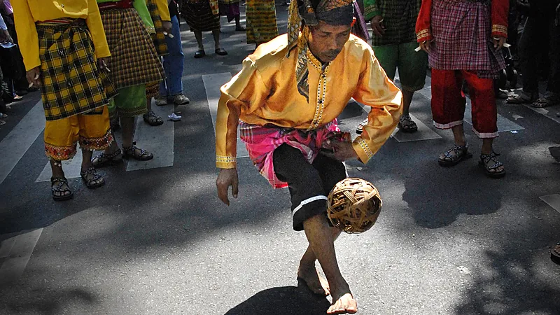 Dua Perunggu Sepak Takraw dan Peran Sulsel di Asian Games Seorang lelaki berbaju kuning bersulam dan bersongkok menimang bola rotan dengan kaki, dikelilingi pemain lain bersarung di atas jalan beraspal
