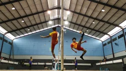 Dua pemain sepak takraw melompat di sisi jaring dalam gelanggang beratap seng, salah satunya melepas tendangan salto ke arah bola