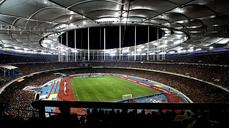 Tribune Stadion Nasional Bukit Jalil penuh penonton pada malam hari dengan lampu sorot menyala di atas lapangan