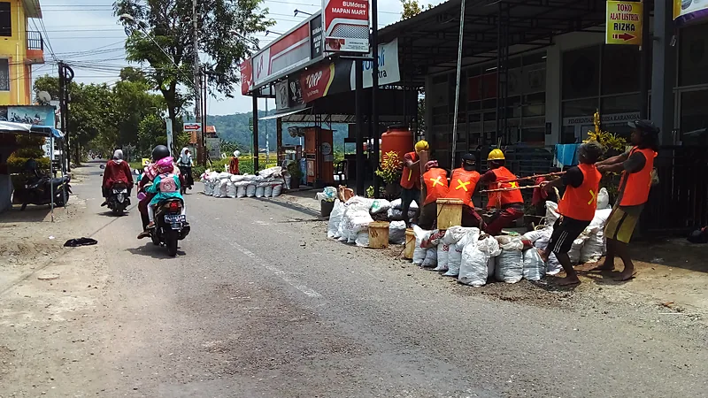 Pekerja berompi oranye dan berhelm kuning menarik tali di tepi jalan di samping tumpukan karung, sementara pengendara sepeda motor melintas