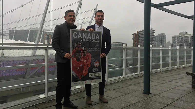 John Herdman berjas gelap memegang poster laga Kanada bersama seorang pemain di teras stadion Vancouver
