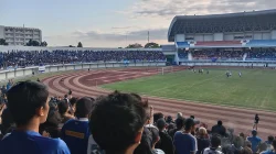 Suasana pertandingan sepak bola di Stadion Mandala Krida Yogyakarta menjelang senja, dengan tribun penuh penonton berbaju biru dan lintasan atletik mengelilingi lapangan