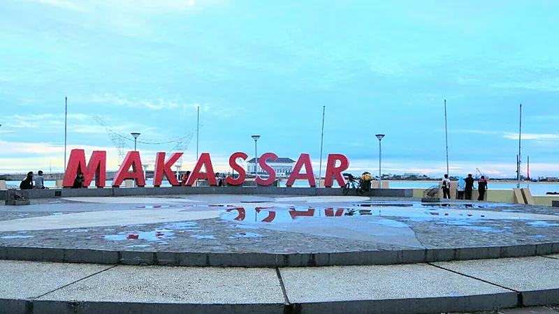 Huruf merah besar bertuliskan MAKASSAR di pelataran tepi laut menjelang senja, beberapa warga duduk dan berjalan di sekitarnya, genangan air memantulkan huruf itu