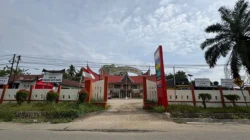 Gerbang kantor pemerintahan desa beratap merah bergaya adat dengan papan nama dan bendera merah putih di halaman