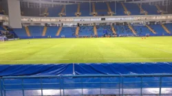 Lapangan hijau sebuah stadion sepak bola malam hari dengan tribun biru bertingkat dan pemain berlatih di kejauhan