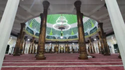 Interior masjid berkubah hijau dengan tiang-tiang berukir keemasan, lampu gantung besar, dan karpet merah bergaris saf