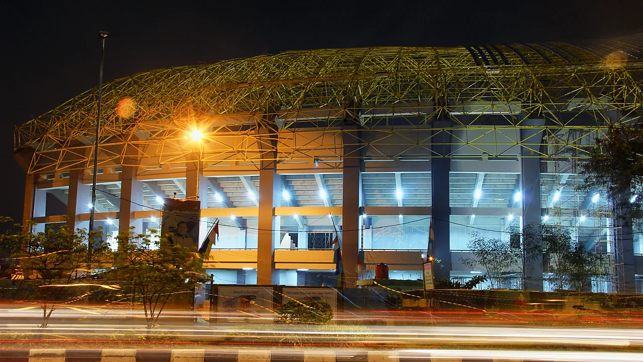 Fasad luar stadion pada malam hari dengan rangka atap baja dan lampu menyala