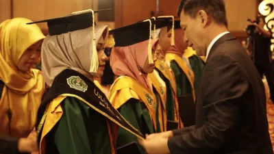Deretan wisudawan berjubah hijau dan toga hitam menerima map dari seorang pria berjas di dalam gedung