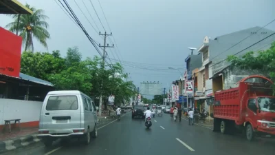 Jalan beraspal di kota Bantaeng dengan deretan ruko, tiang listrik, dan tugu bertuliskan Bulukumba Berlayar di kejauhan