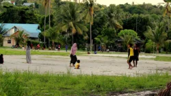 Lapangan sepak bola kampung berlatar pohon kelapa dengan warga menonton dan anak-anak bermain di pinggir lapangan