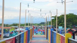 Jalan setapak berpagar warna-warni di taman rekreasi tepi pantai dengan untaian lampu di atasnya, deretan kursi plastik dan warung di kedua sisi, serta perbukitan di kejauhan