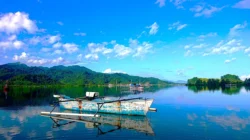 Perahu nelayan bercadik bercat putih biru mengapung di perairan tenang dengan perbukitan hijau di kejauhan