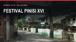 Jalan desa di Tana Beru dengan papan penunjuk bertuliskan Pembuatan Perahu Phinisi dan rumah panggung kayu di kedua sisi, dengan teks judul "Festival Pinisi XVI" dan label Rubrik.co.id - Bulukumba di bagian atas gambar
