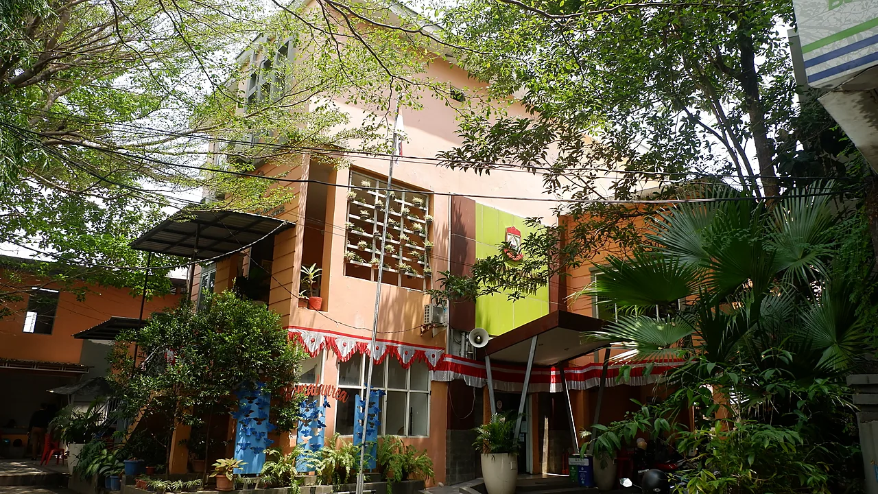 Gedung kantor kecamatan bercat jingga dengan panel hijau bergambar lambang kota dan taman kecil di depannya